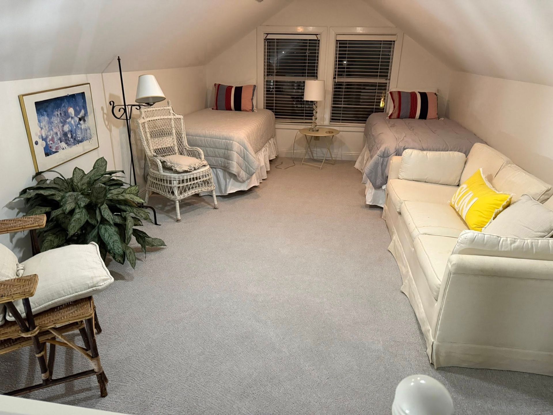 Attic bedroom with two beds, a wicker chair, and a white sofa. Soft gray carpet and walls.
