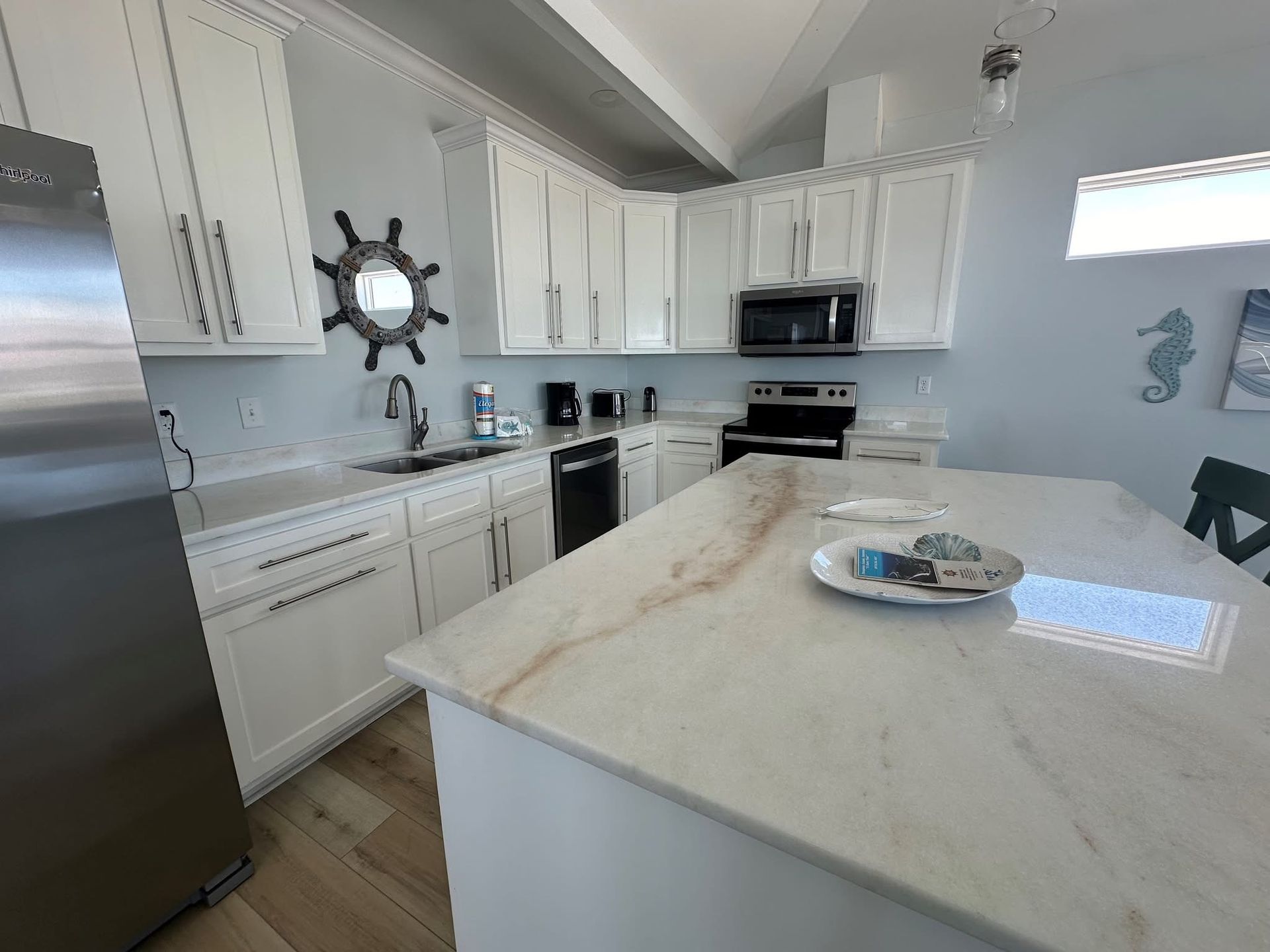 A modern white kitchen with a large marble island, stainless steel refrigerator, and nautical decor.