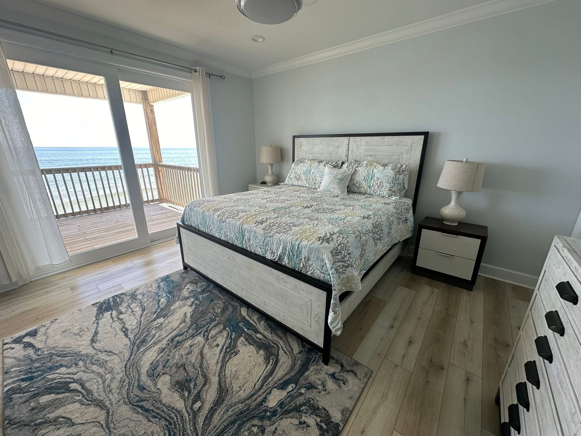 A bedroom with a queen bed, blue and white patterned bedding, a matching rug, and a balcony overlooking the ocean.
