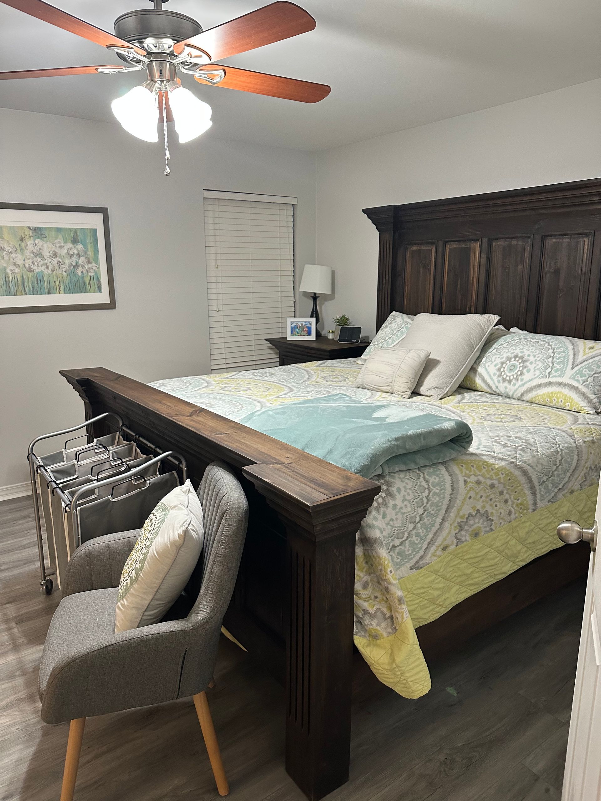 A bedroom featuring a dark wood bed with patterned bedding, a gray armchair, and a ceiling fan.