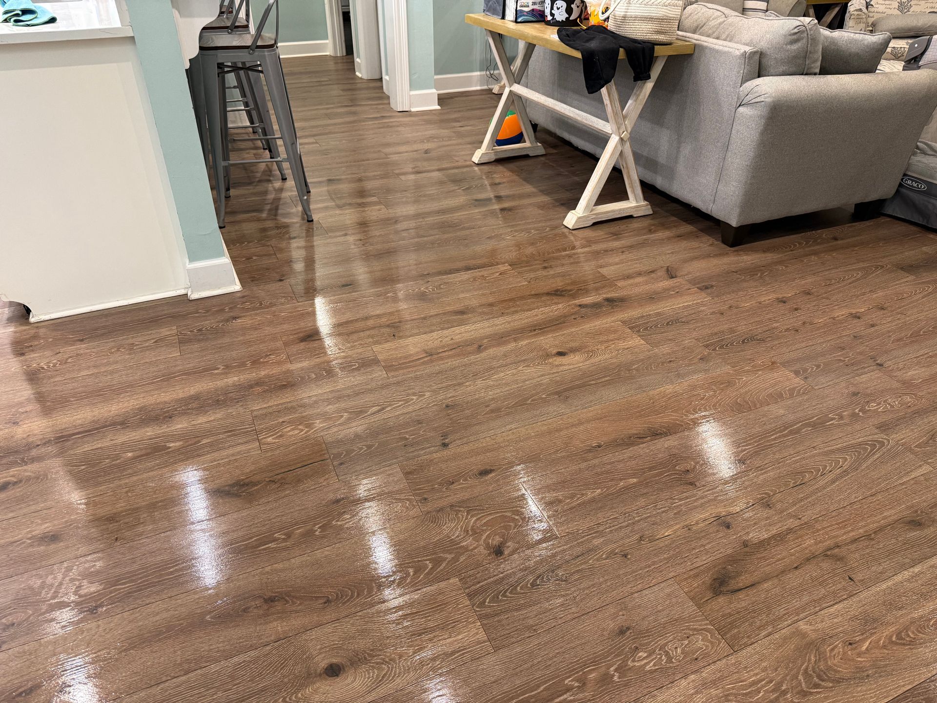 Interior view of a room with polished brown wood-look flooring, a kitchen counter with bar stools, and a gray sofa.