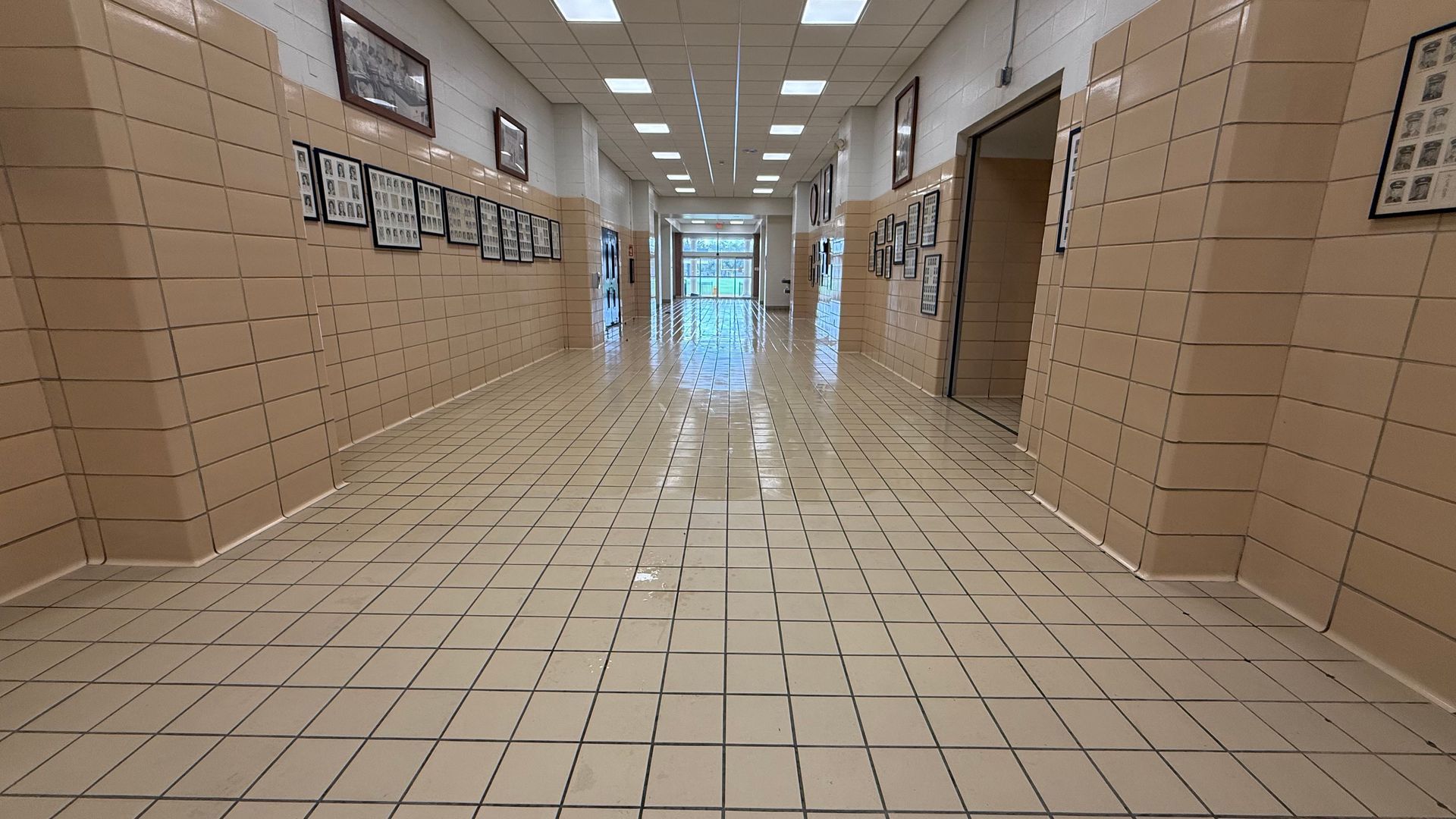 A long, empty school hallway with beige tiled walls and floors, illuminated by recessed ceiling lights.