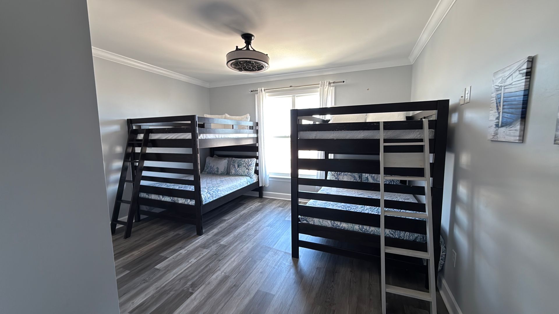 A bedroom with two dark wooden bunk beds, gray flooring, light walls, and a window with curtains in the center.