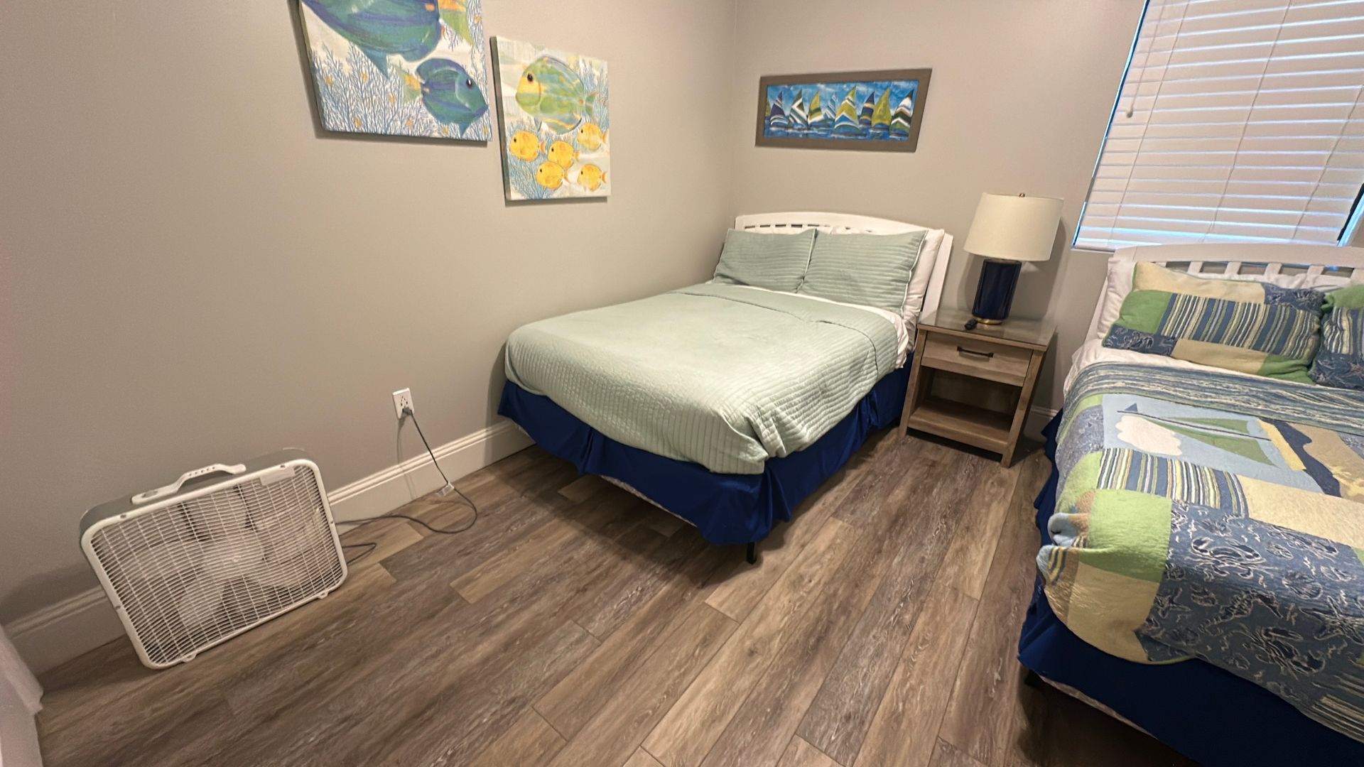 A bedroom with two beds, a nightstand, and a box fan on the wood floor, decorated with coastal artwork on the walls.