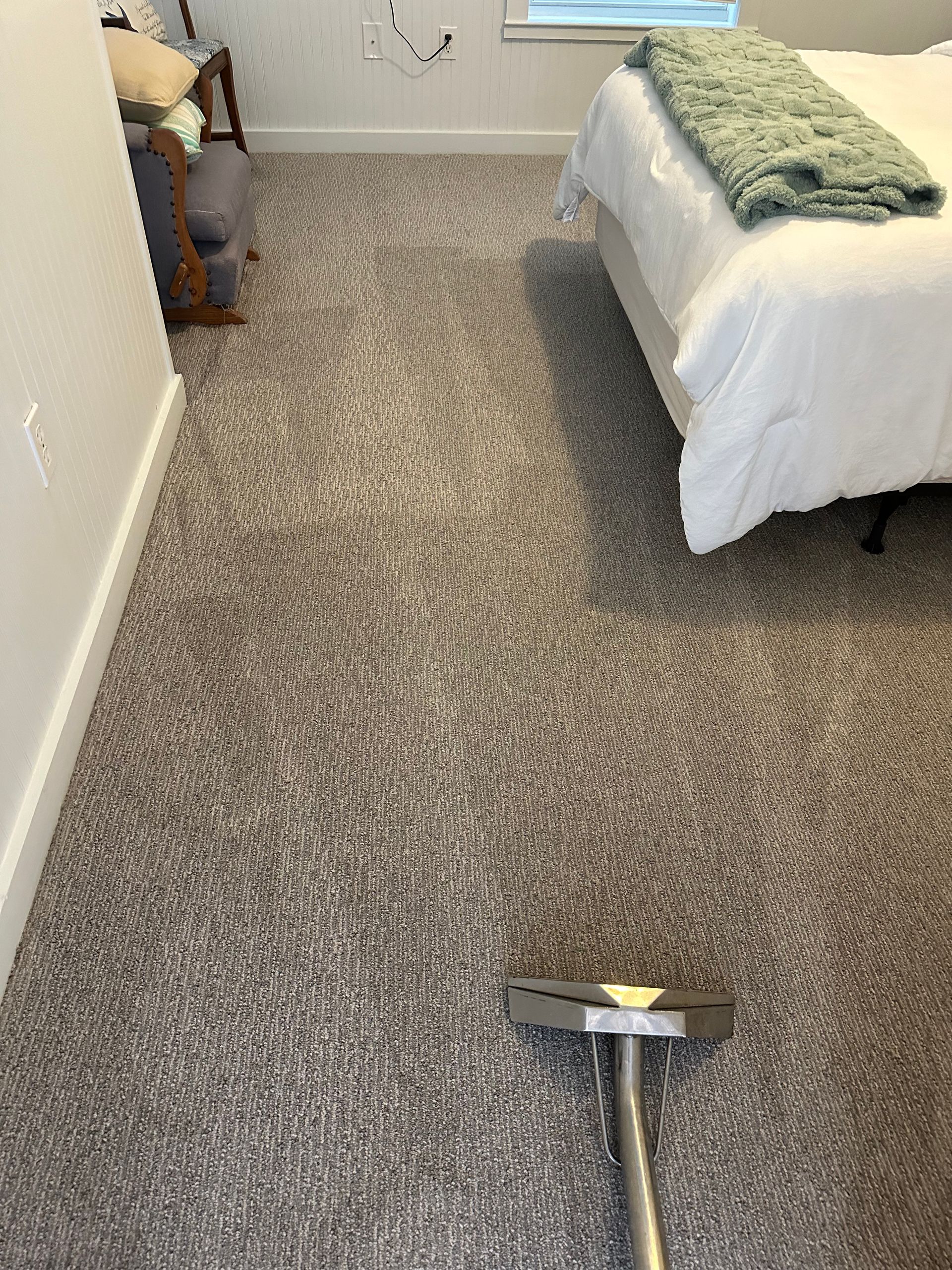 A vacuum cleaning attachment rests on a gray patterned carpet in a bedroom with a bed and chair nearby.