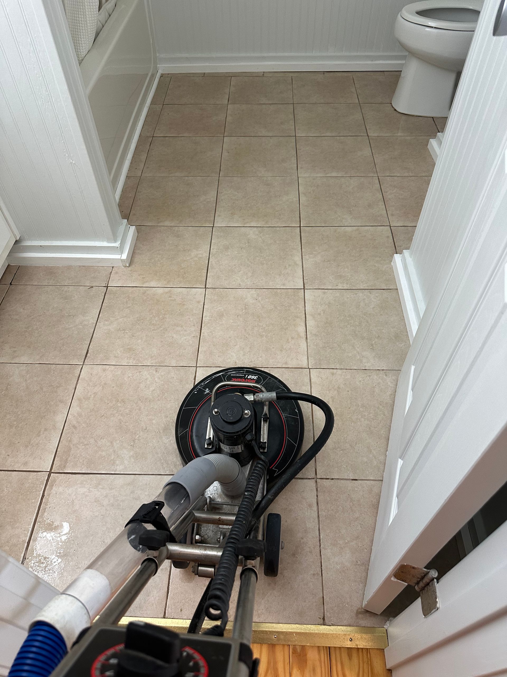 A commercial floor scrubber cleans beige tile in a bathroom near a bathtub and toilet.