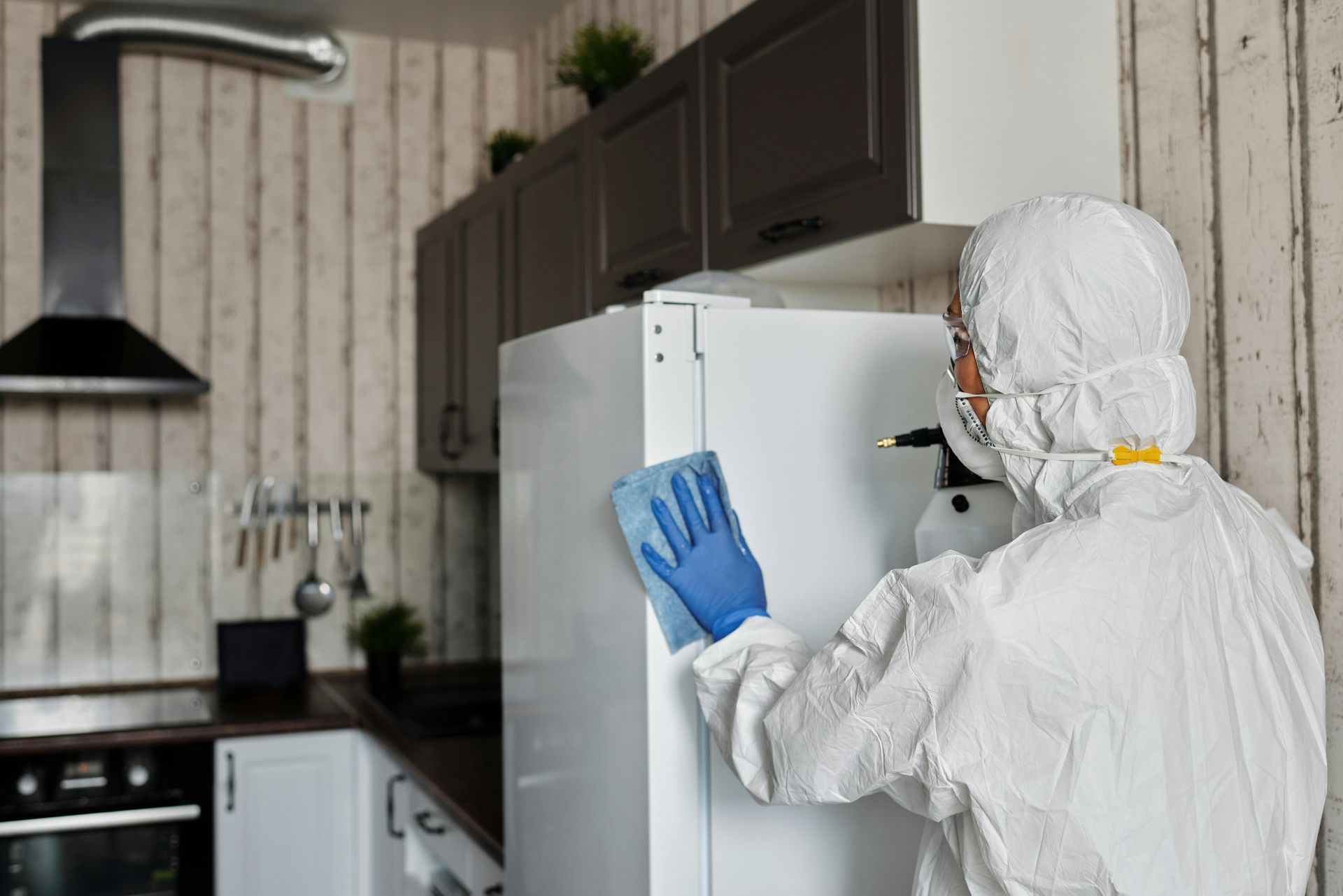 Person in hazmat suit sanitizing a white refrigerator in a kitchen with gray cabinets.