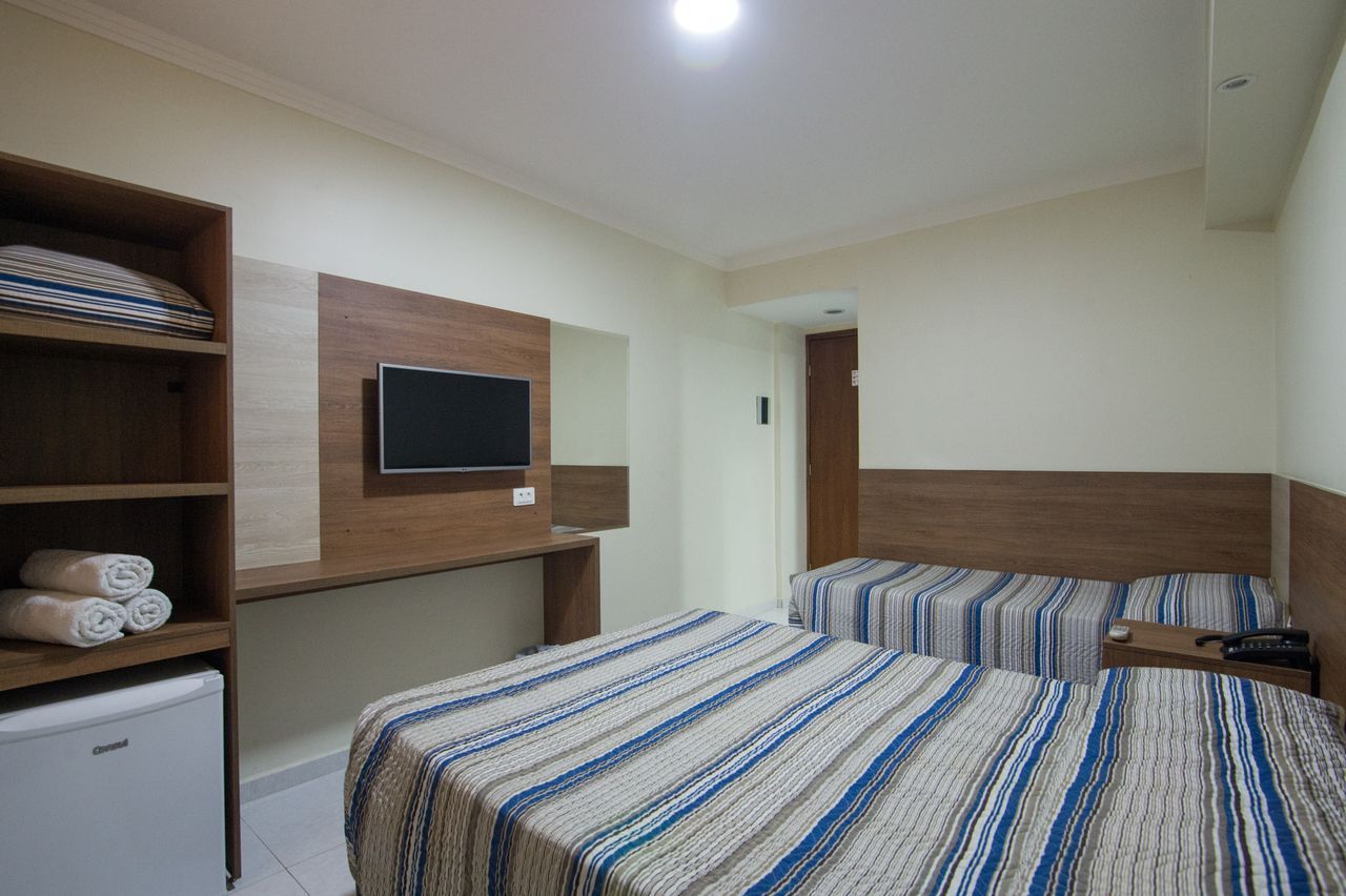 Quarto de hotel com duas camas, TV, frigobar e armário. Decoração em marrom e branco, roupa de cama listrada.