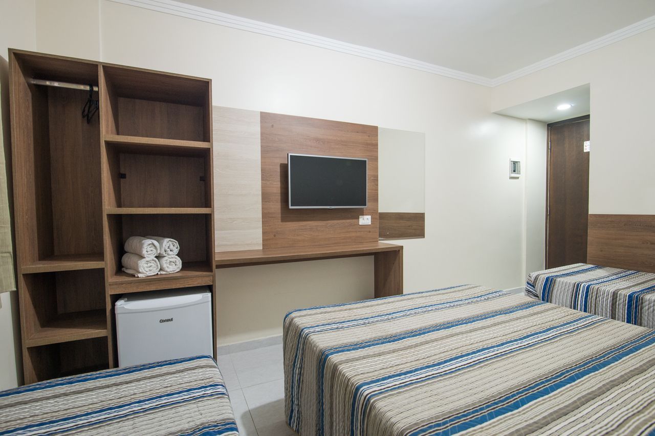 Quarto de hotel com duas camas, guarda-roupa, frigobar e TV na parede; cores neutras.