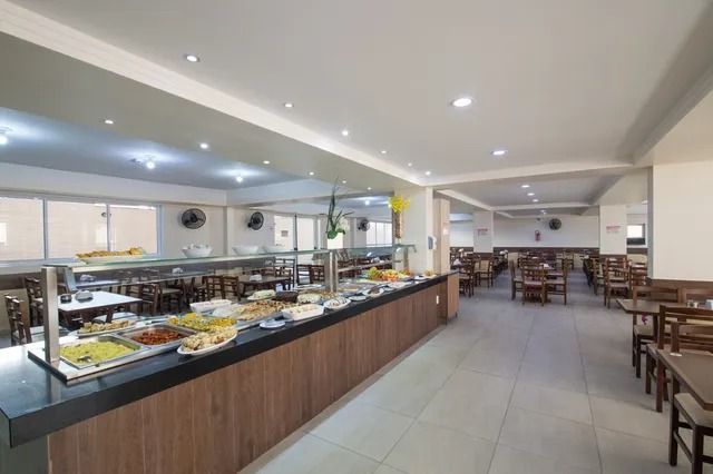 Restaurante em estilo bufê com comidas expostas. Fileiras de mesas com cadeiras estendem-se até o fundo.