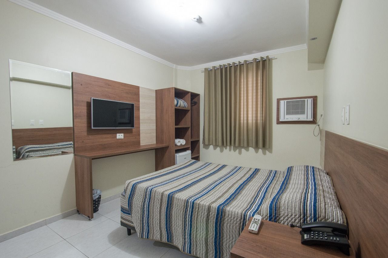 Quarto de hotel com cama, escrivaninha, TV, janela com cortina, armário e frigobar.