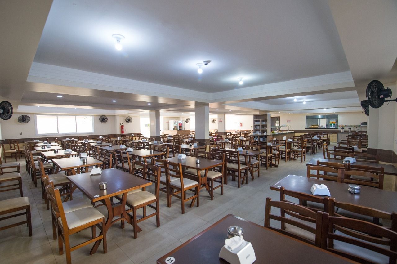 Sala de jantar vazia com muitas mesas e cadeiras de madeira. Paredes brancas, luminárias no teto.