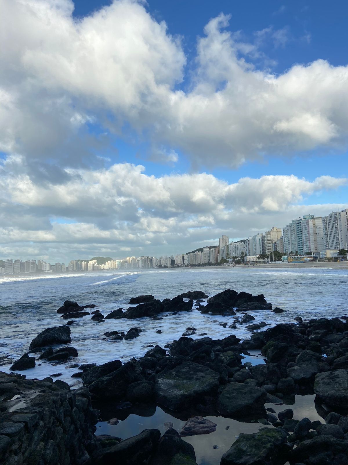 Costa rochosa e oceano com ondas brancas, prédios da cidade sob um céu azul nublado.