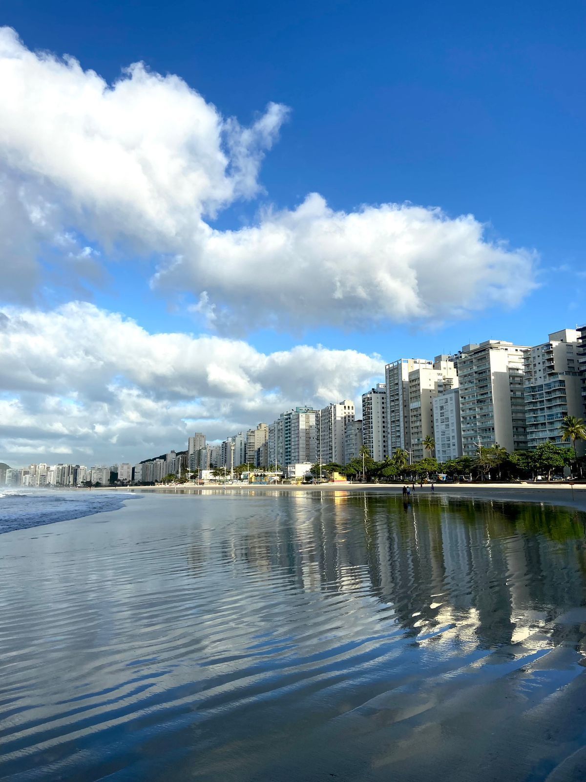 Praia com águas calmas refletindo edifícios sob um céu azul com nuvens brancas e fofas.