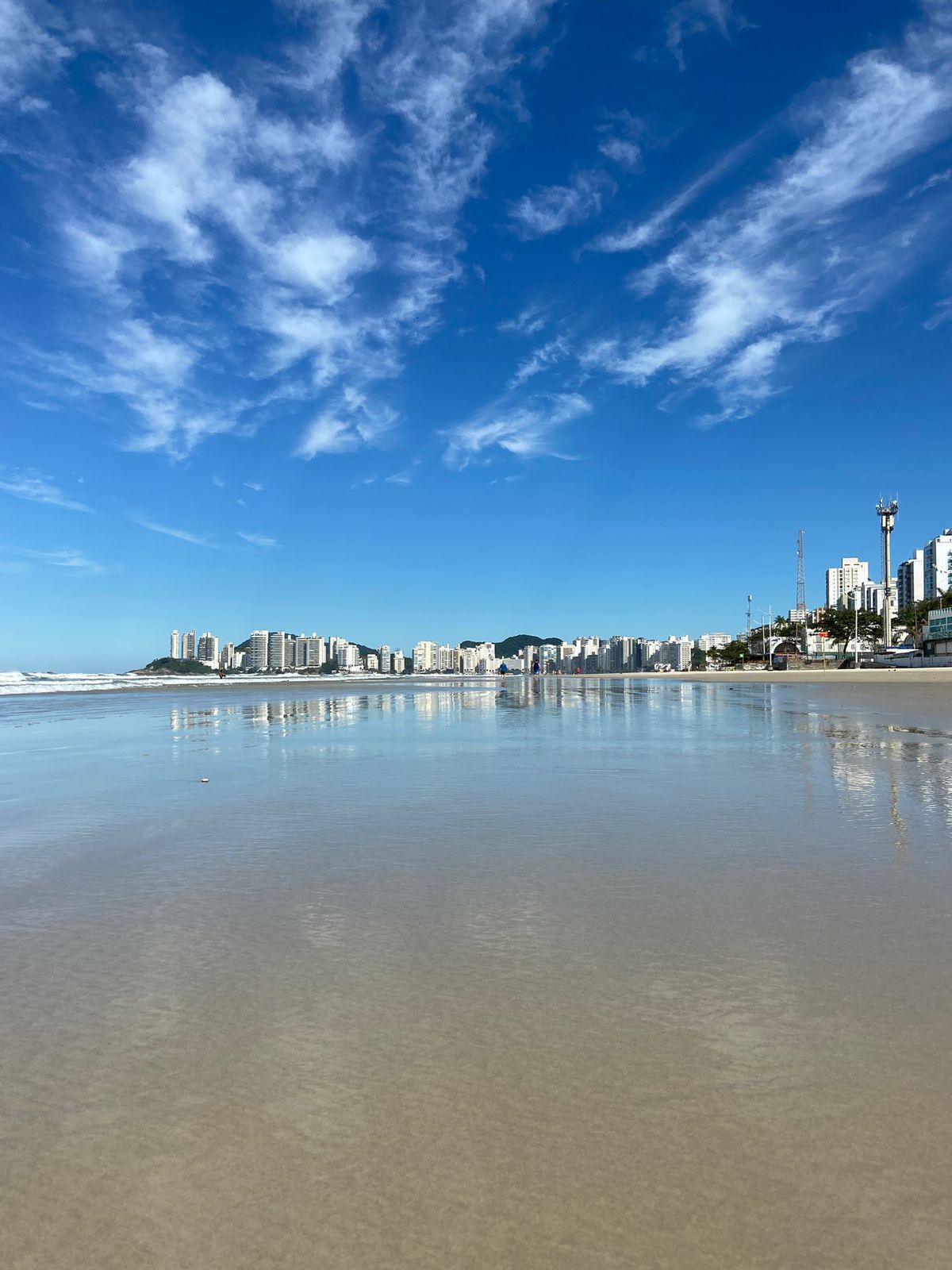 Praia com prédios e céu azul nublado refletido na areia molhada.