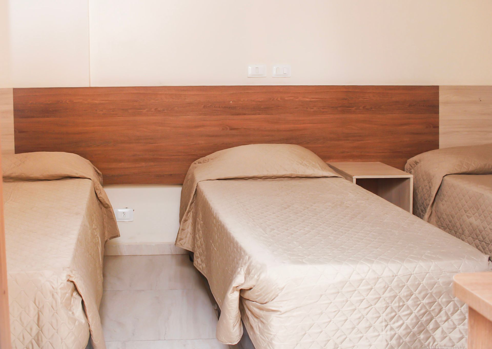 Três camas de solteiro em um quarto de hotel, com roupa de cama bege e cabeceira de madeira.