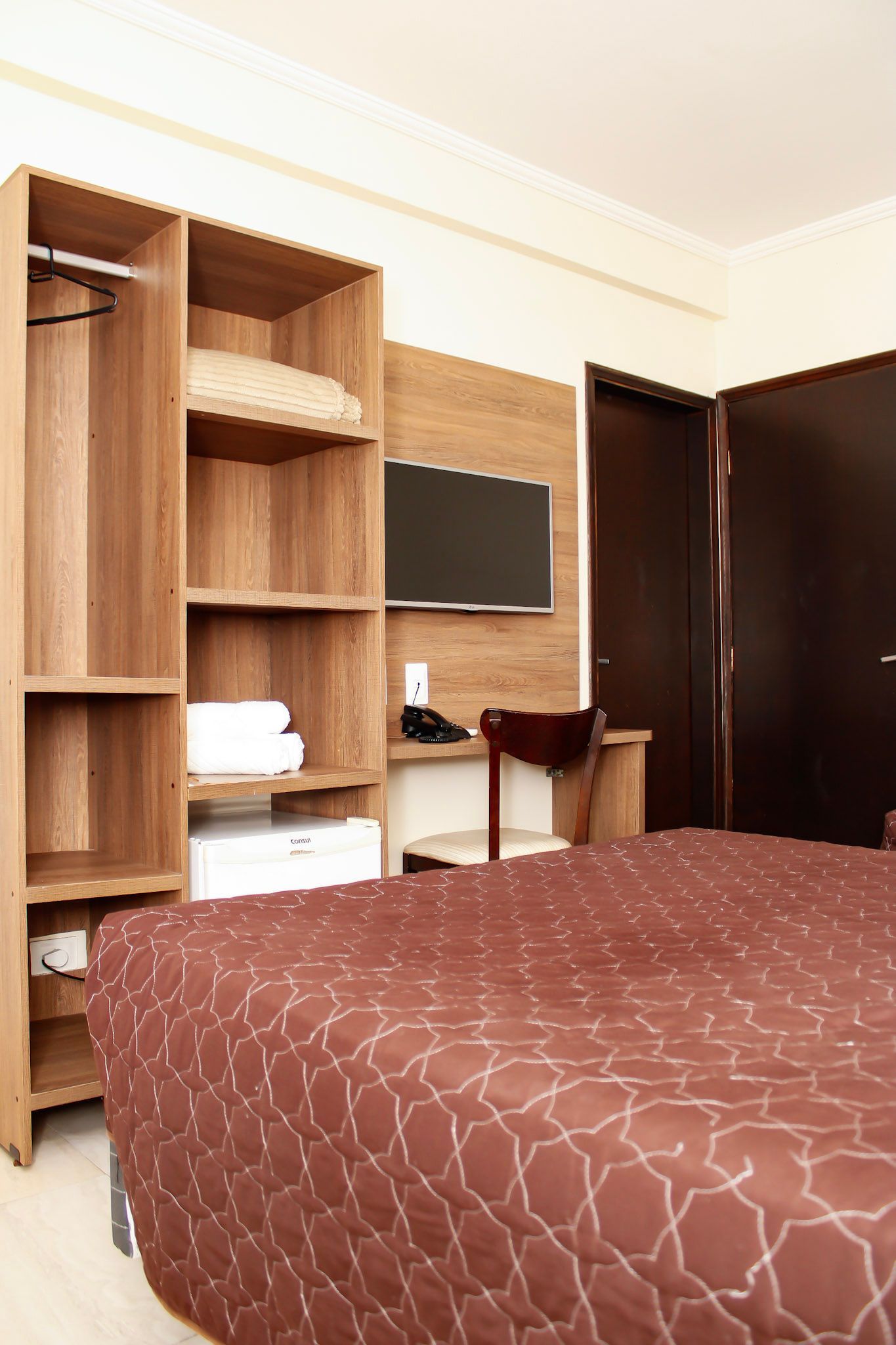 Quarto com guarda-roupa de madeira, TV, colcha marrom e porta marrom fechada.