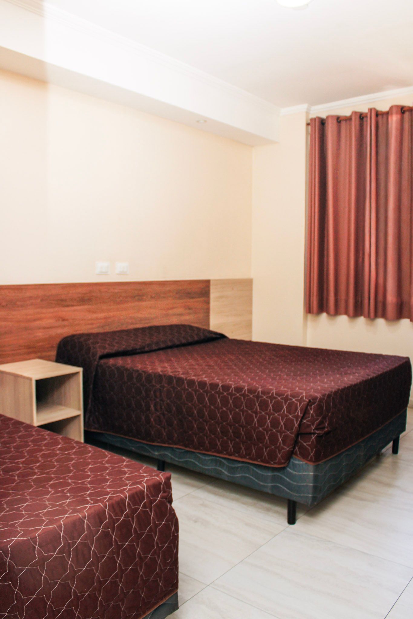 Quarto de hotel com duas camas, colchas estampadas em marrom, cortinas marrons, paredes claras e um criado-mudo.