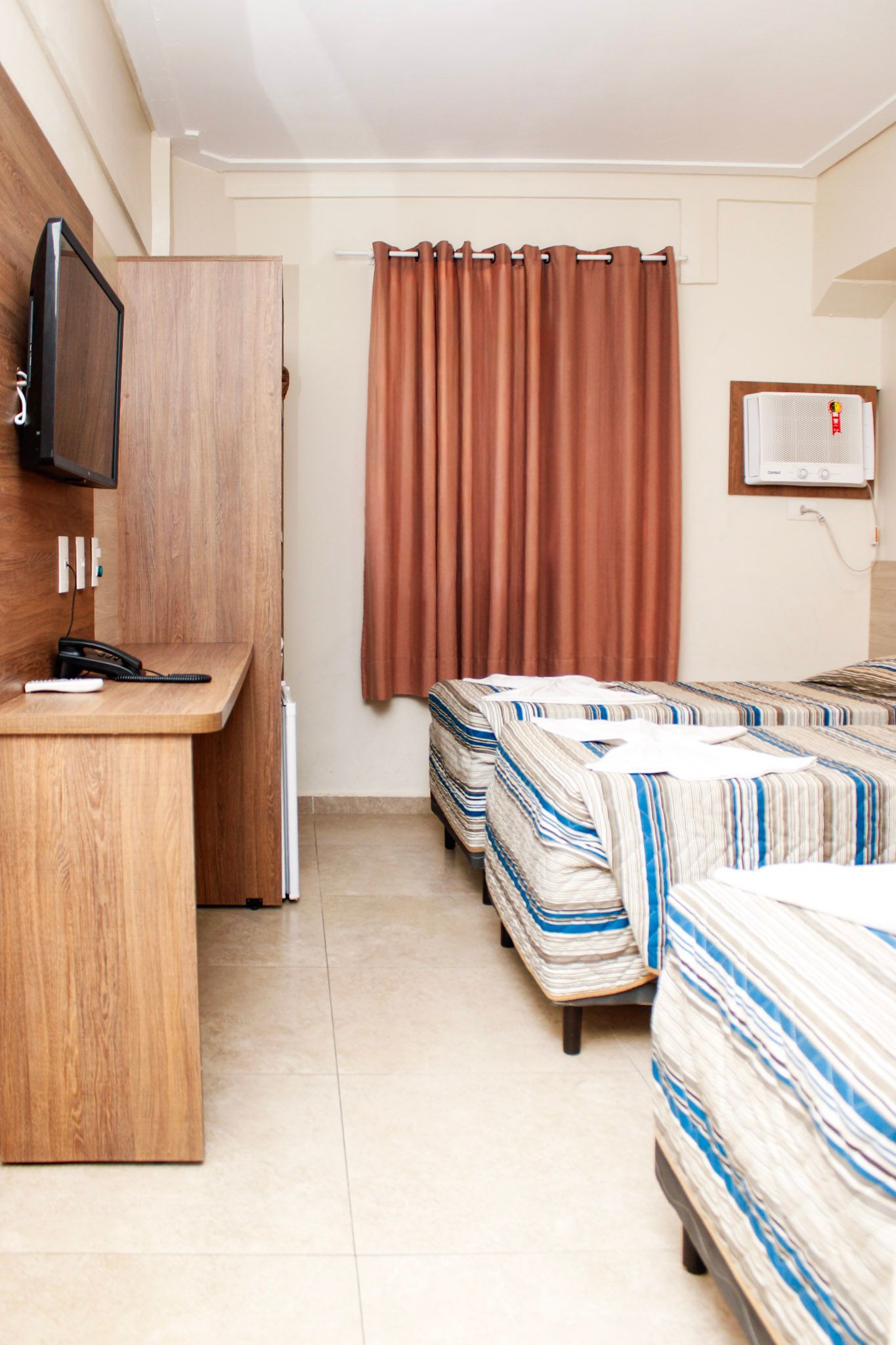 Quarto de hotel com três camas, uma TV, uma escrivaninha e cortinas marrons.