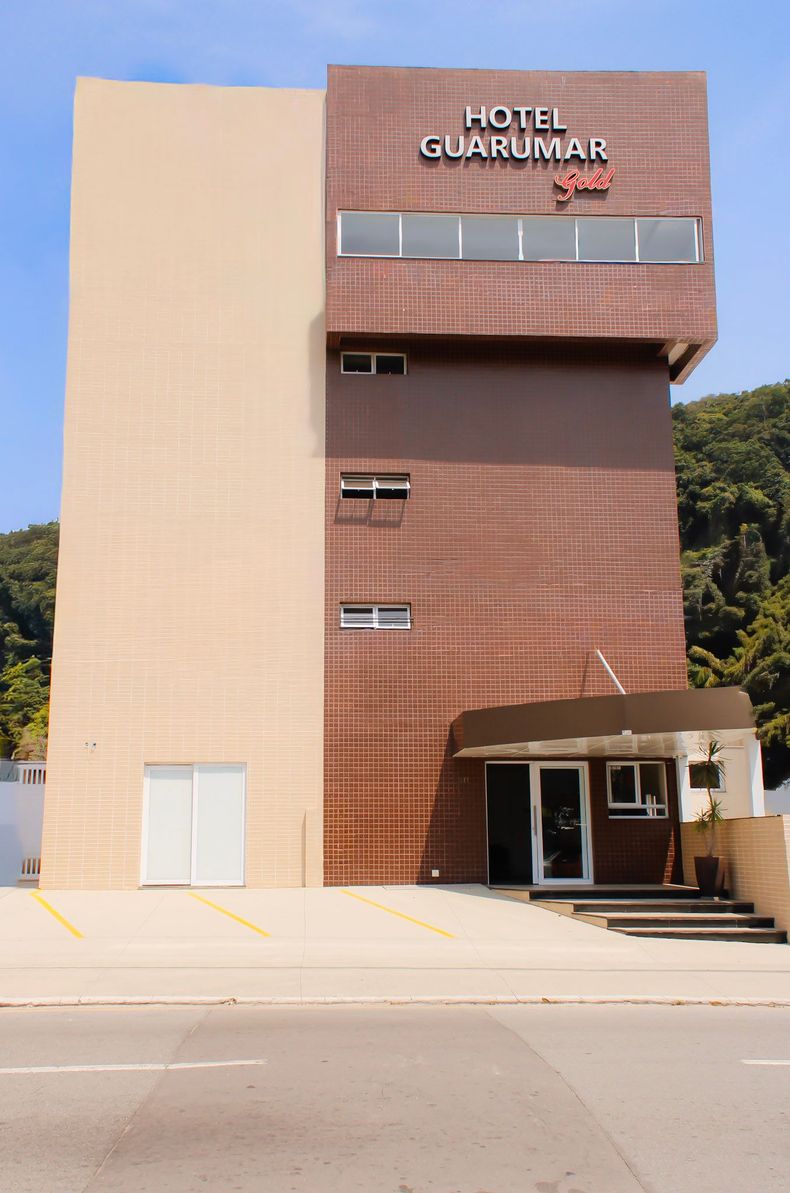 Prédio do Hotel Guarujá com fachada bege e marrom, entrada de vidro e placa no topo.