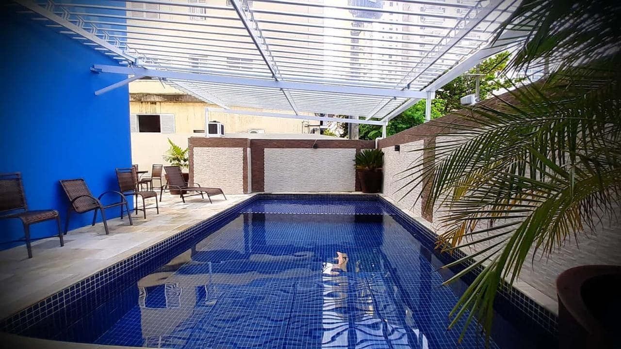 Piscina com palmeiras, água azul e prédios ao fundo. Dia ensolarado.