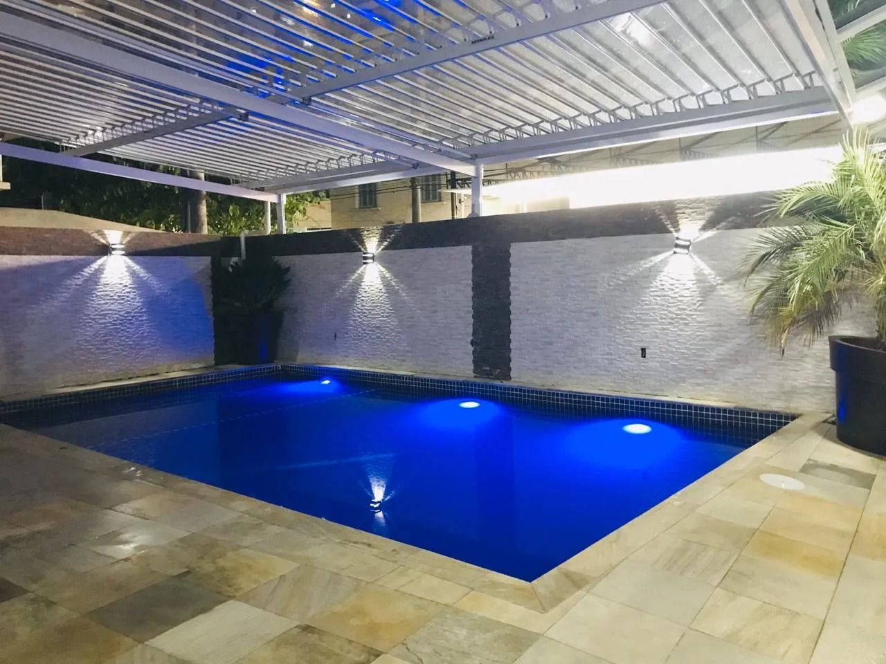 Piscina iluminada sob uma pérgola branca, com parede texturizada em branco e preto, pátio de pedra e plantas em vasos.