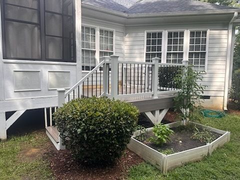 White deck with railing, steps, and screened porch next to a white house, with a bush and garden.