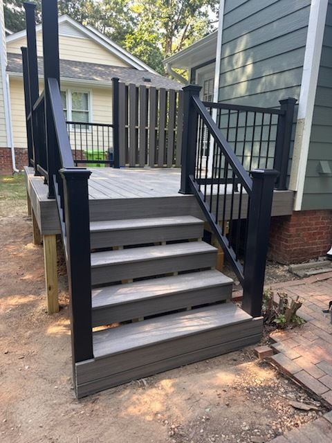 Gray composite deck with black railings and stairs, leading to a house in a yard.