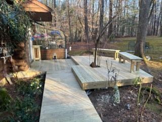 Wooden deck and walkway in a wooded area. Tree in deck center, benches, and small greenhouse visible.