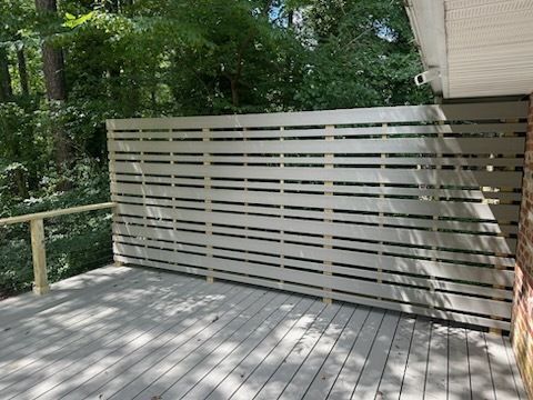 Gray horizontal slat fence on a wooden deck, beside a brick wall, with trees in the background.