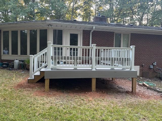 White deck with railing attached to a brick house, in a yard with trees.