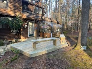 A wooden deck with a bench extends from a brown house with a greenhouse, in a wooded area.