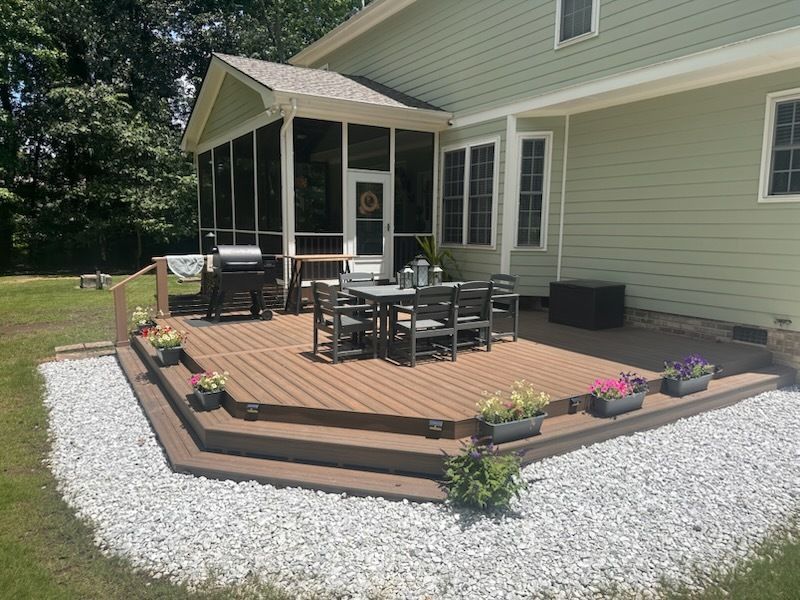 Backyard deck with seating, grill, and planters, next to a green house with screened porch and gravel border.