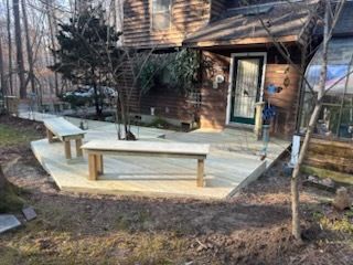 Wooden deck with benches near a house with a door. The scene is outdoors with trees and dirt.
