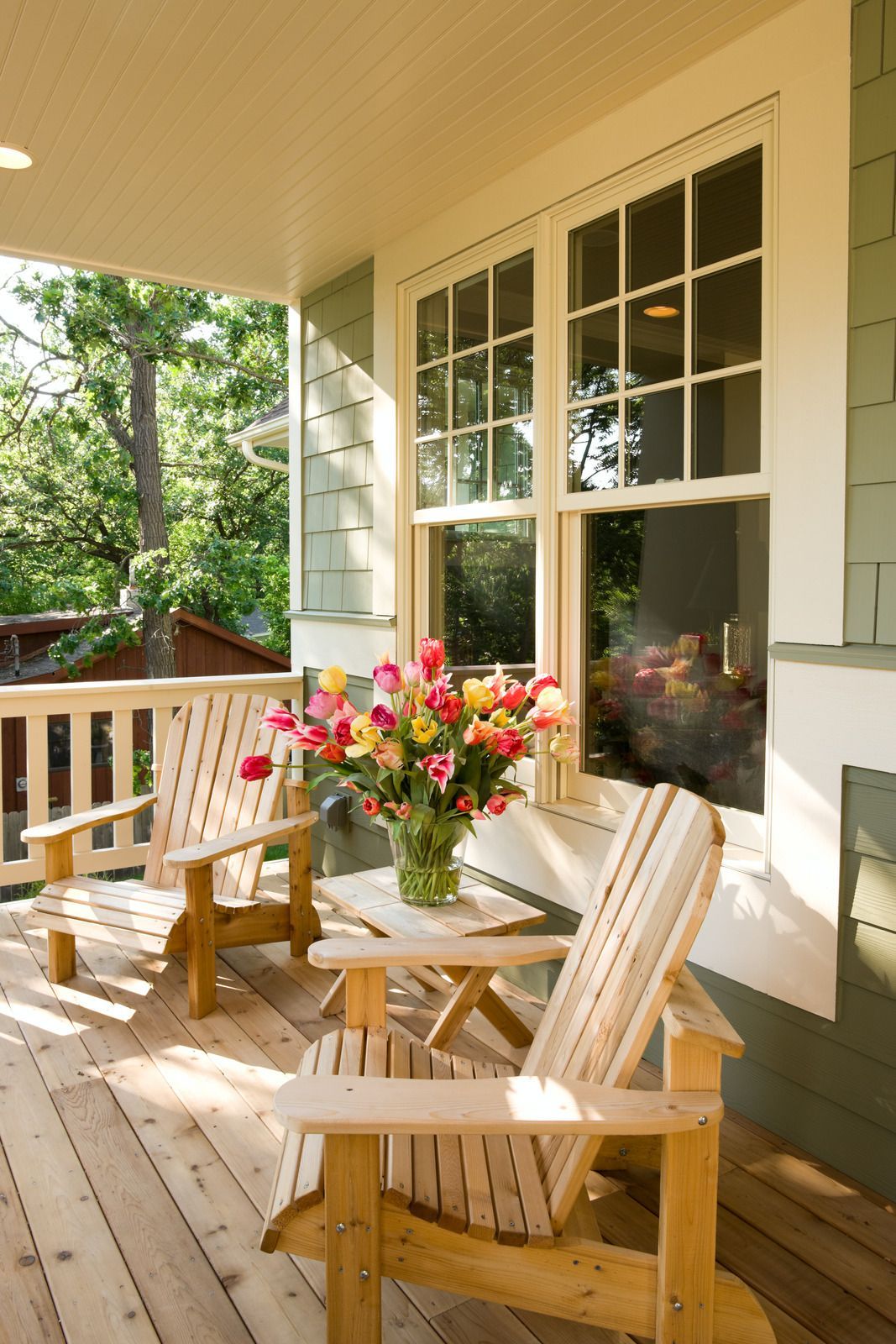Two wooden Adirondack chairs and small table with flowers on a covered porch.