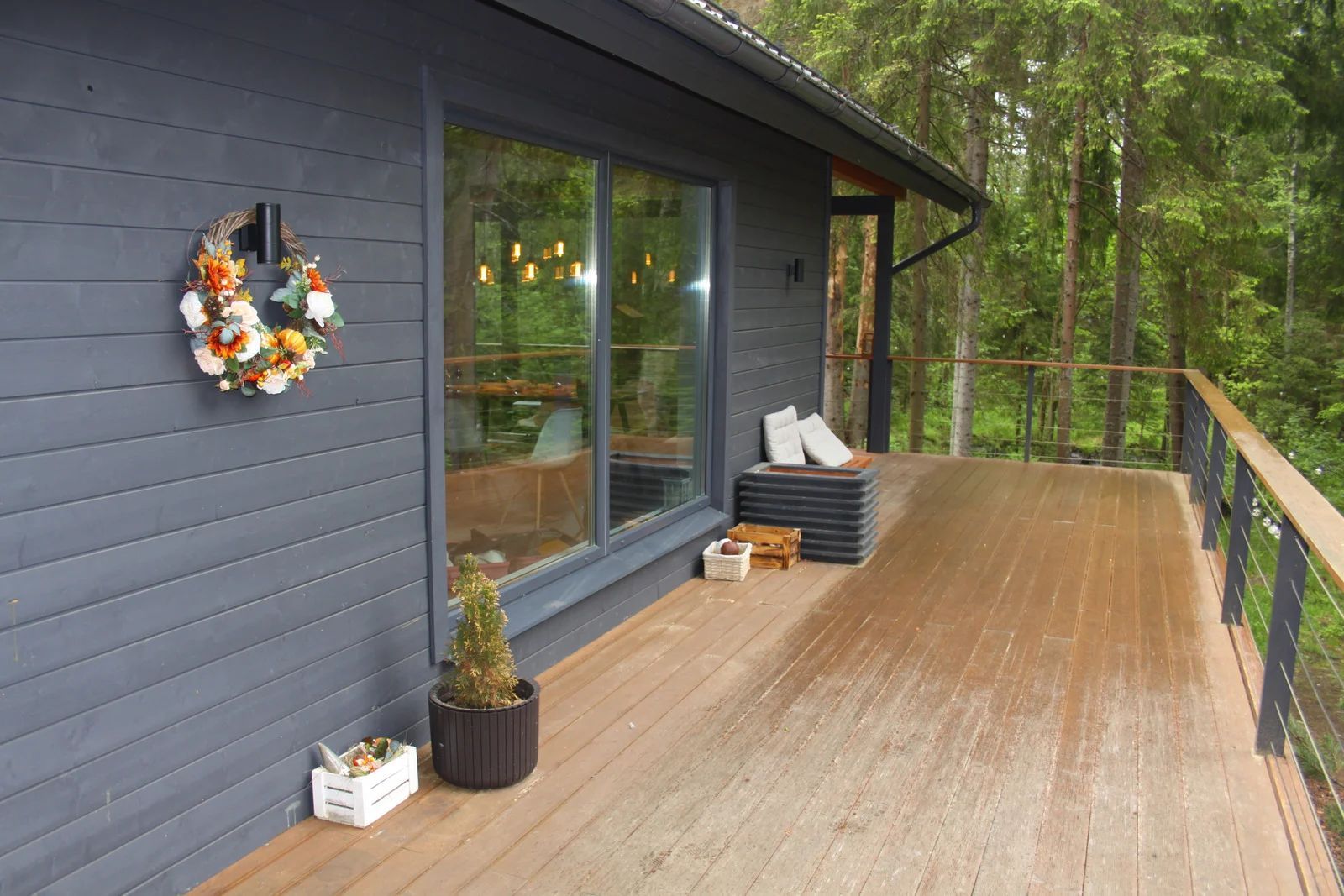 A wooden deck attached to a house with dark gray horizontal siding, a floral wreath, and a view of a forest.