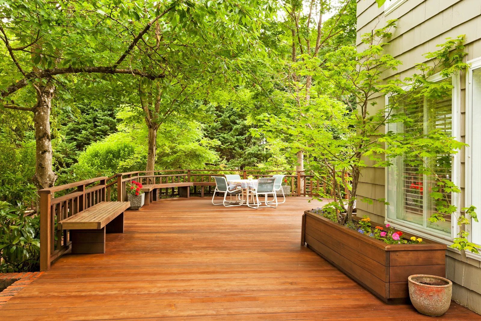 Wooden deck with trees, bench, table, and chairs, adjacent to a house with a window.