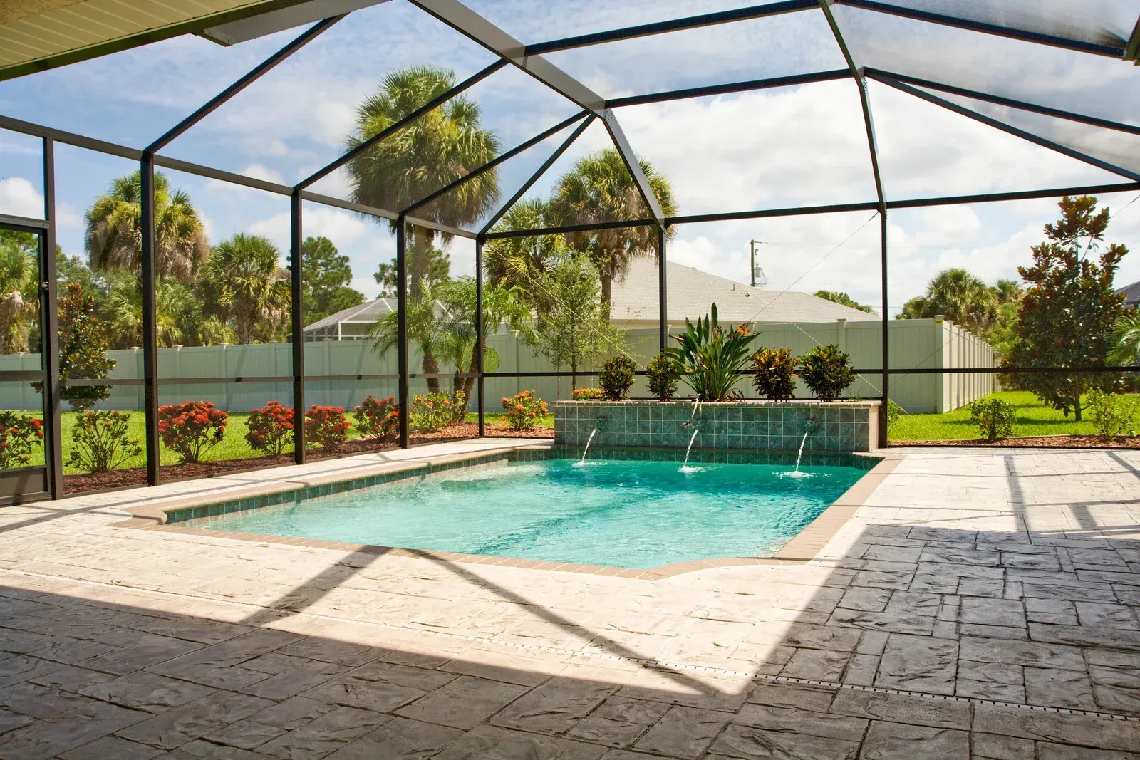 Pool enclosure with a rectangular pool, water fountains, and plants in a sunny outdoor setting.