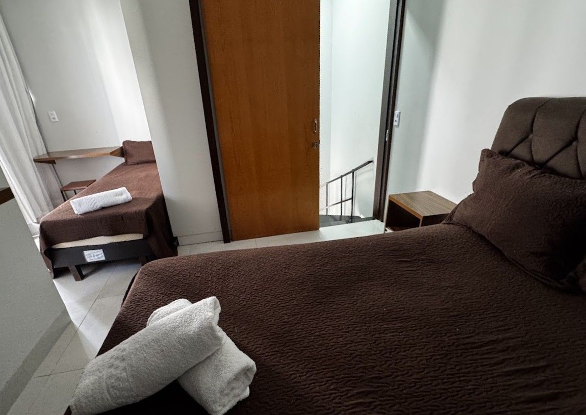 Duas camas marrons em um quarto, uma delas com toalhas enroladas. Uma porta e uma escada são visíveis.