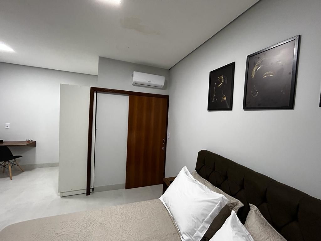 Quarto com cama, sofá marrom escuro, paredes cinza claro, duas obras de arte emolduradas, porta de madeira, escrivaninha ao fundo.