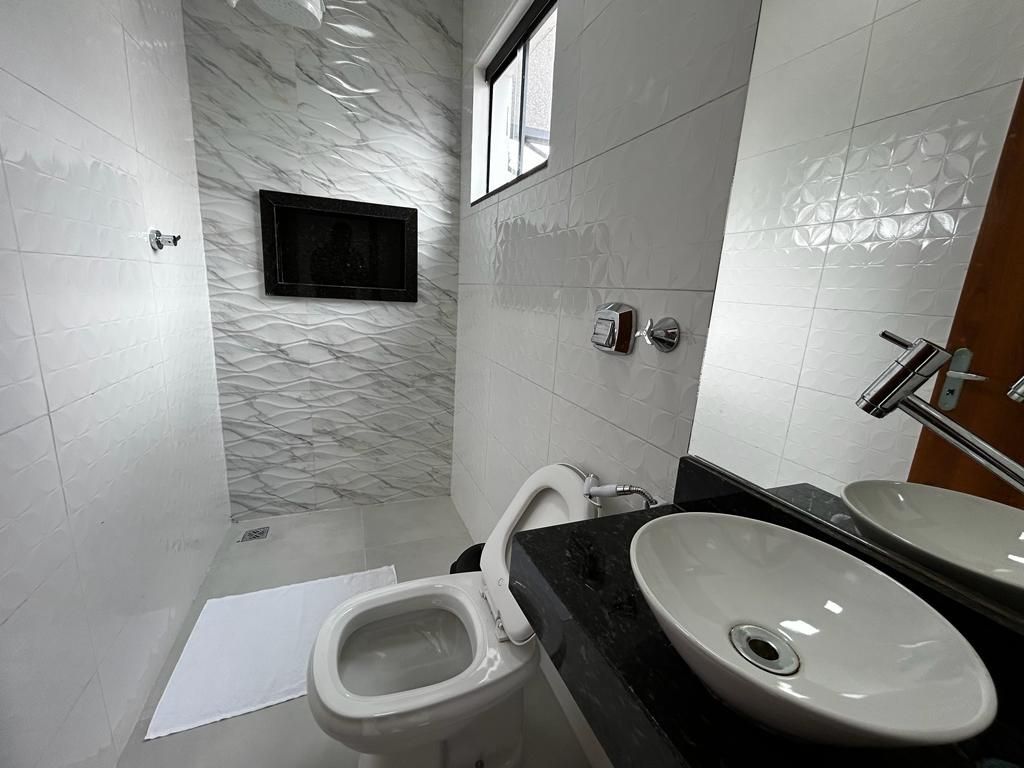 Banheiro moderno com paredes de azulejos brancos, vaso sanitário, pia e uma pequena TV.