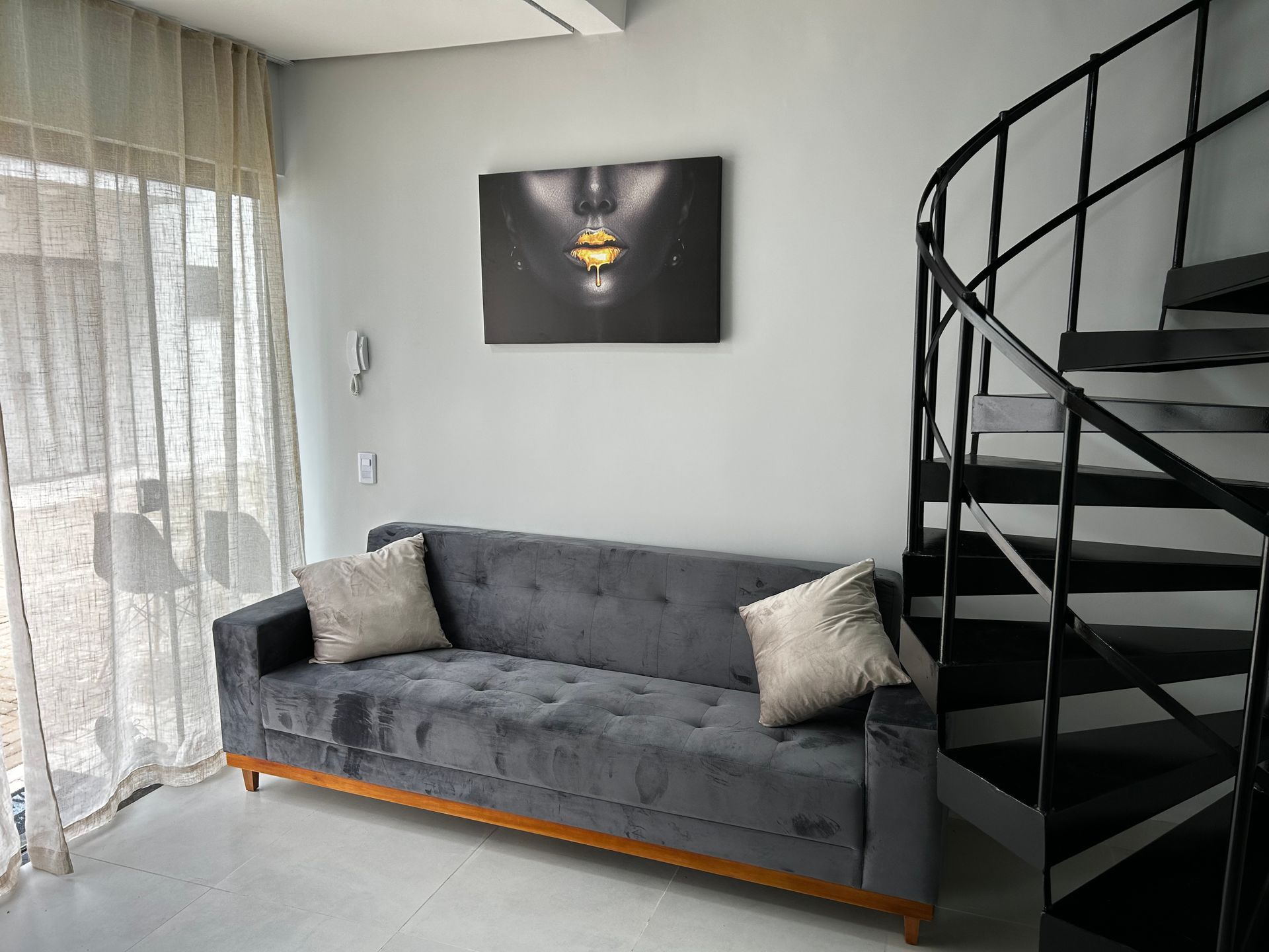 Sofá cinza com almofadas, obra de arte e escada em espiral em uma sala com janela.