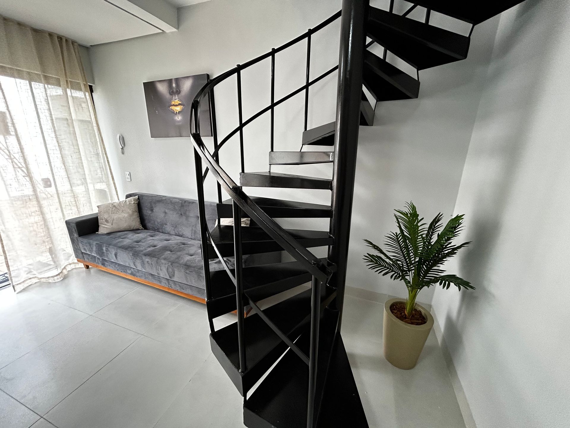 Escada em espiral preta em um interior branco moderno, ao lado de um sofá e uma planta em vaso.
