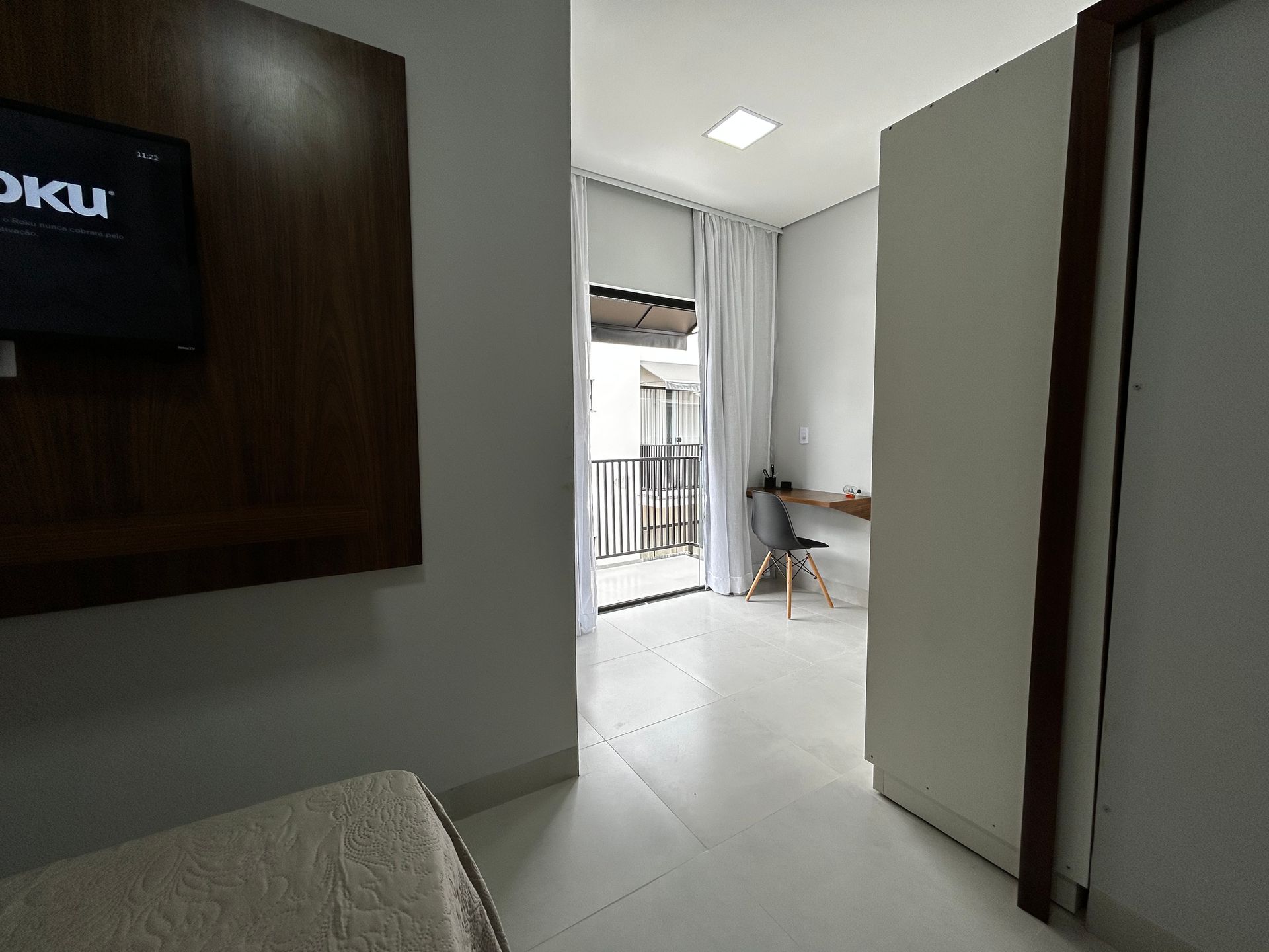 Corredor com TV e varanda que leva a uma escrivaninha e cadeira. Cores claras e neutras predominam.