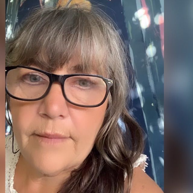 Mujer con gafas y flequillo canoso, mirando a la cámara. Lleva una blusa blanca con un fondo de cortina estampada.