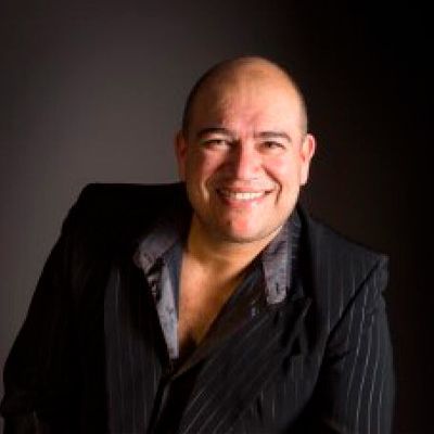 Hombre con traje oscuro sonriendo, inclinado ligeramente hacia adelante contra un fondo oscuro.