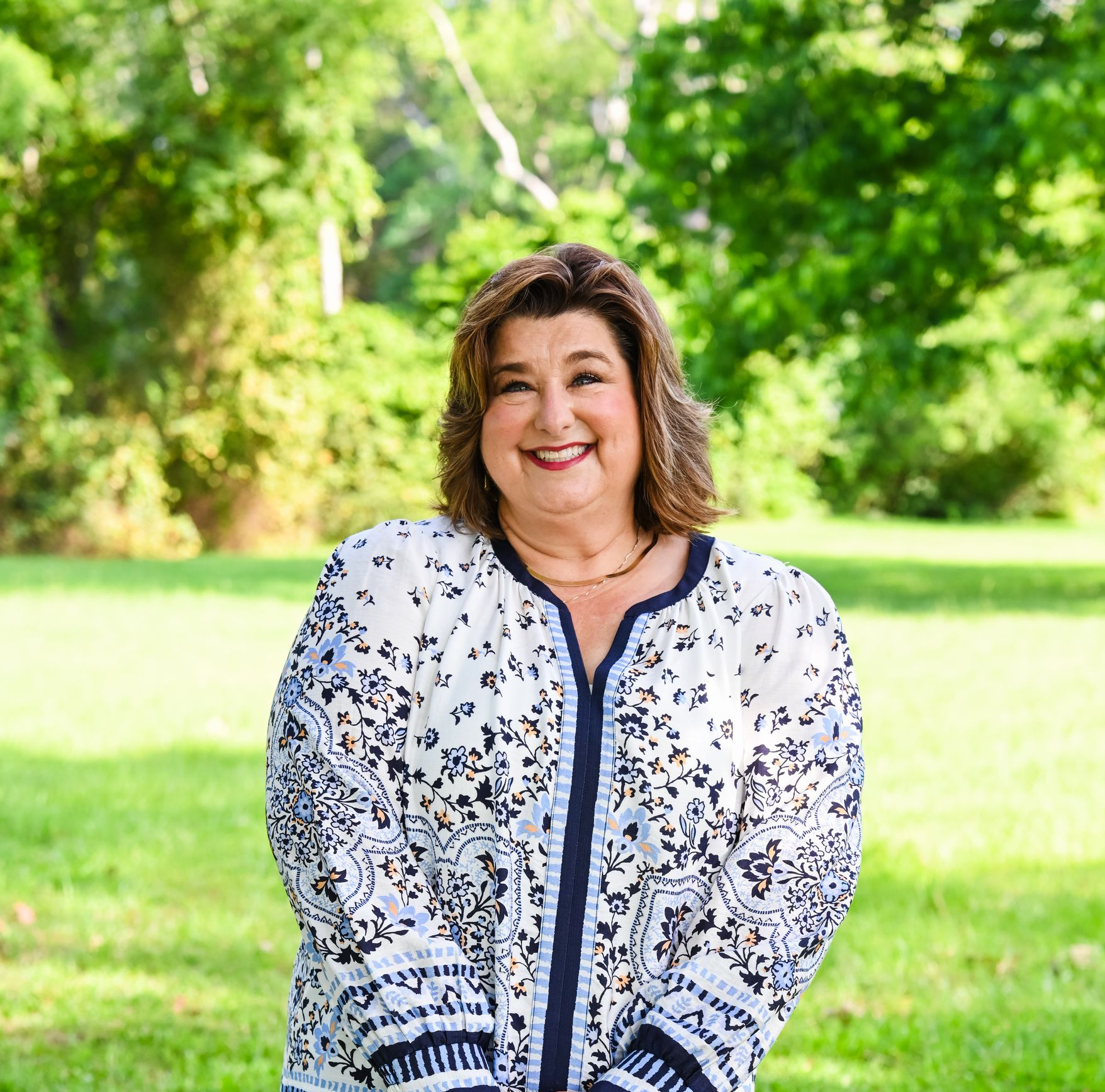 Smiling woman in a white floral blouse standing outdoors in a sunny green park