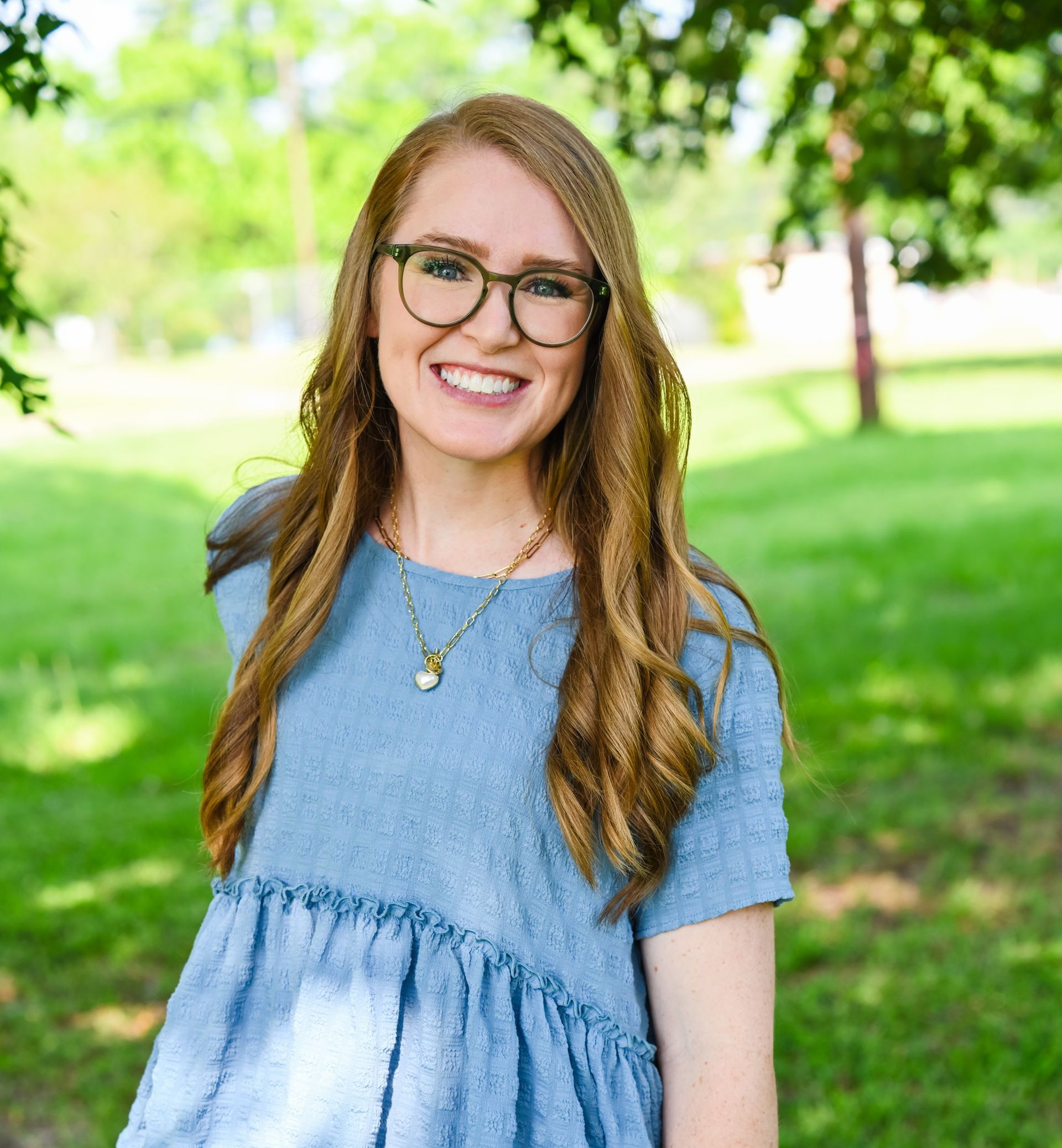 Smiling woman with glasses in a blue dress standing outdoors in a green park