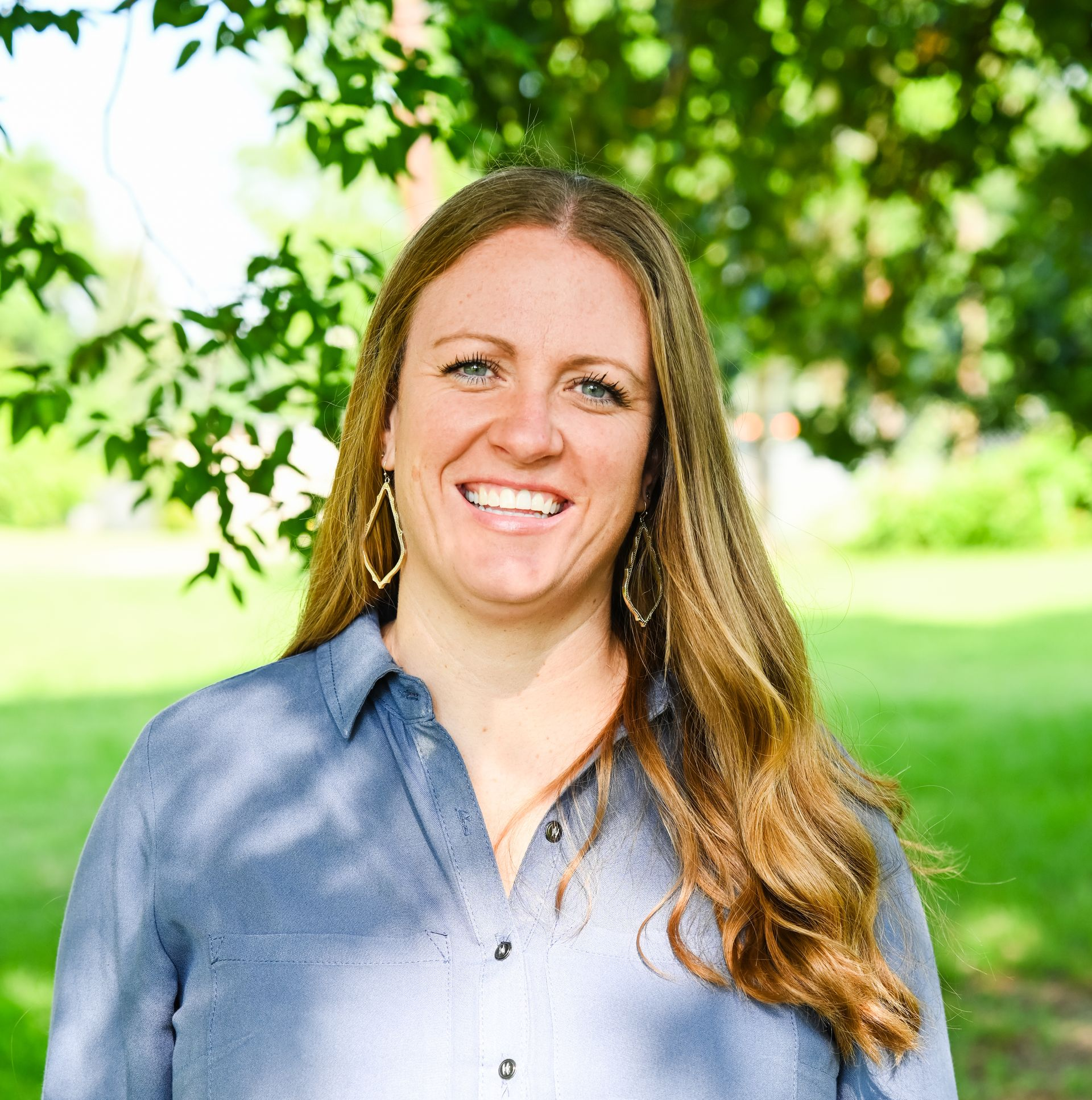 Smiling person in a blue shirt standing outdoors with green trees and grass in the background