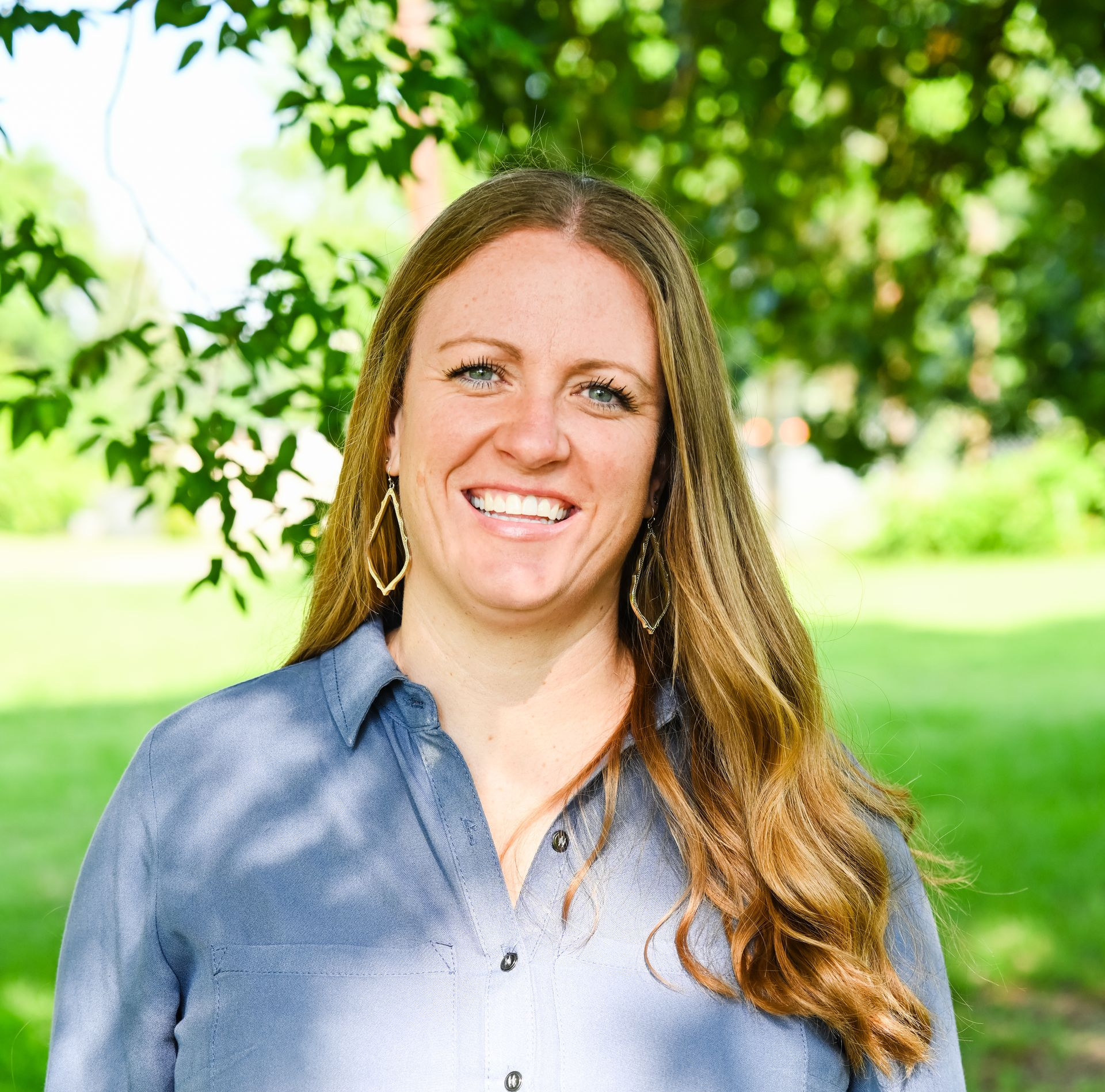 Smiling person with long hair in a blue shirt standing outdoors in a green park.